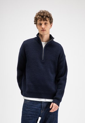 Young man with curly hair wearing a navy blue half-zip sweater over a white shirt and dark blue jeans, standing against a plain background.