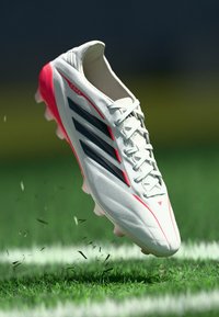 Λευκό αθλητικό παπούτσι με ροζ και μαύρες λεπτομέρειες, κομψό σχέδιο, υφή στο επάνω μέρος και ελαφρύ πέλμα, ανυψωμένο πάνω από το γρασίδι.