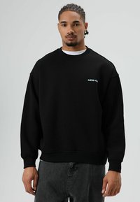 Sweat-shirt noir à col rond, coupe ample et poignets côtelés. Présente un petit logo bleu « RAÍCES Vivas » sur la poitrine. Texture de matière douce.