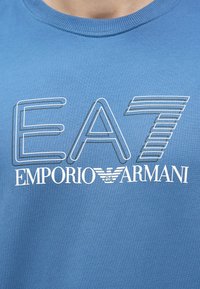 Zila kokvilnas T-krekls ar baltu "EA7 EMPORIO ARMANI" logotipu baltā kontūrā un spārnotu emblēmu. Mīksta tekstūra, parasts piegriezums.