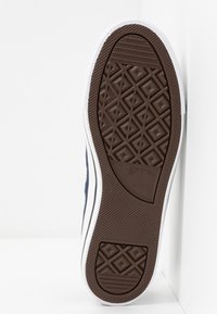 Hnědá gumová podrážka tenisky se geometrickým vzorem. Obsahuje bílé lemování a logo v zaoblené oblasti. Pozadí je bílé.