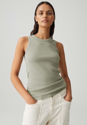 Femme aux longs cheveux foncés portant un haut sans manches vert clair côtelé et un pantalon blanc, debout les mains dans les poches.