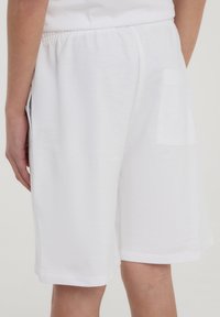Pantaloni corti bianchi in cotone con vita elasticizzata, tasche laterali e posteriori, vestibilità rilassata e texture liscia. I bordi sono finiti in modo semplice.
