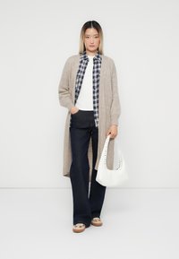Beige gestrickte Strickjacke über einem weißen T-Shirt, kombiniert mit einem karierten Hemd und dunklen Schlagjeans. Weiße Tasche mit Ausschnitten.