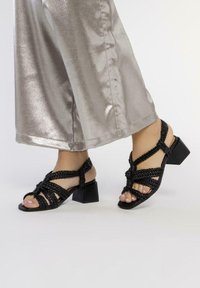 Pies en sandalias negras de tiras trenzadas con tacón cuadrado, llevadas con pantalones anchos plateados brillantes, sobre un fondo liso.