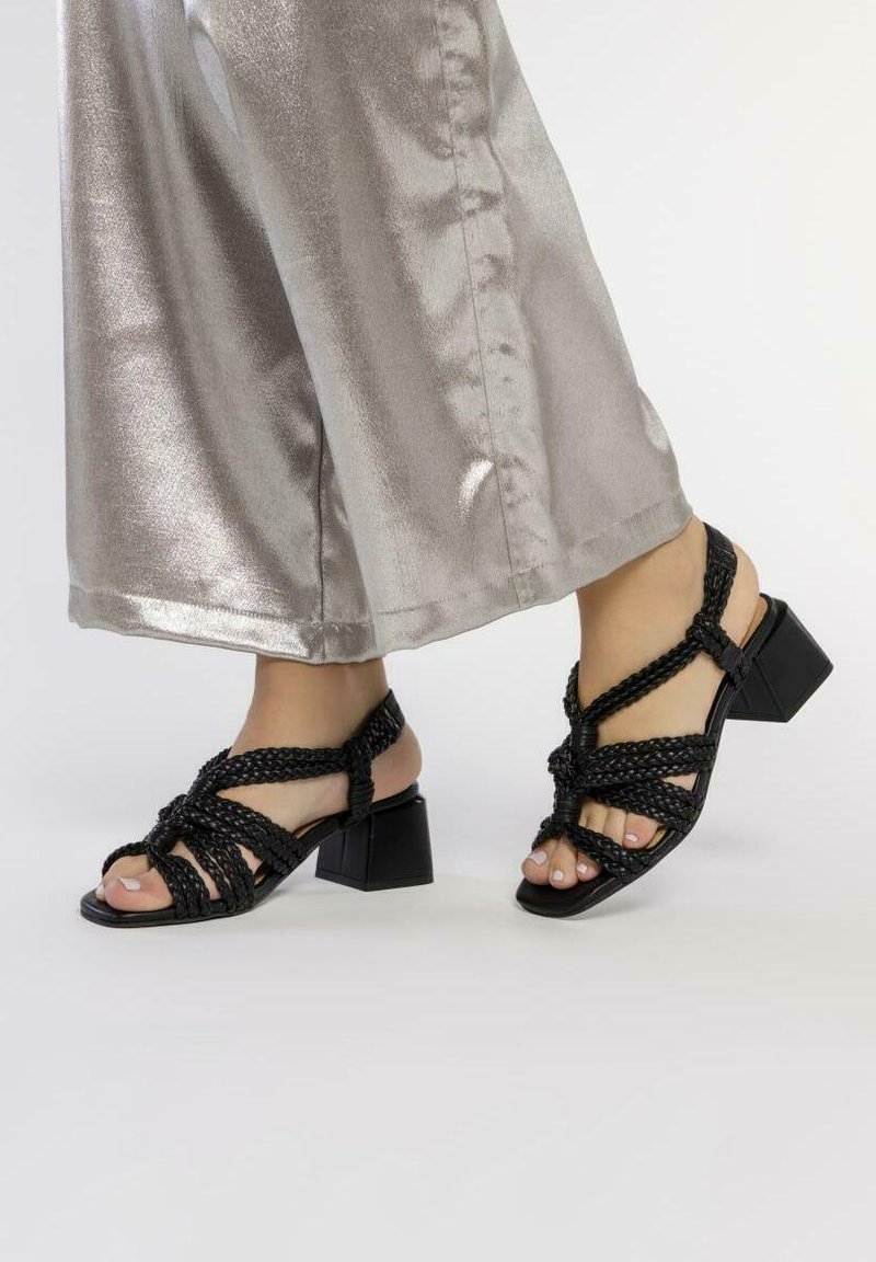 Pies en sandalias negras de tiras trenzadas con tacón cuadrado, llevadas con pantalones anchos plateados brillantes, sobre un fondo liso.