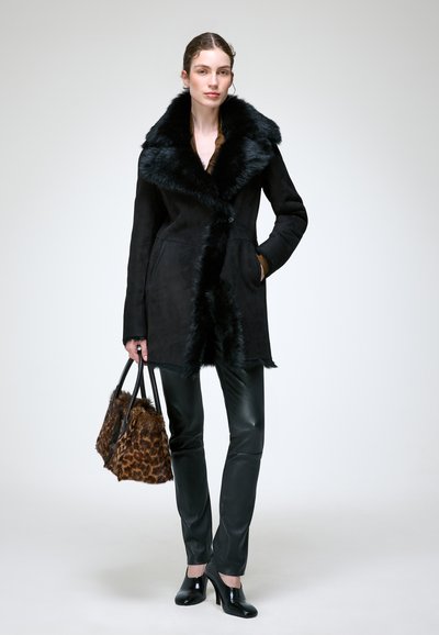 Femme portant un manteau noir bordé de fourrure, un pantalon en cuir noir, des chaussures noires à talons, et tenant un sac à main en fourrure imprimé léopard marron.