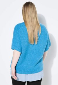 Ljusblå stickad tröja med korta ärmar, med en ribbad nederdel. Bärs över en vit och blå randig skjorta, i kombination med svarta byxor.