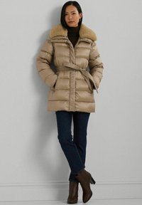 Beige gewatteerde jas met een bontkraag, een ceintuur in de taille en ritszakken, gecombineerd met donkere jeans en bruine enkellaarzen.