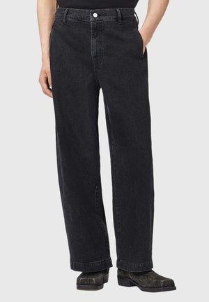 Sorte vidde jeans båret med sorte støvler, hænder i lommerne, vist fra taljen til fødderne på en ensartet lys baggrund.