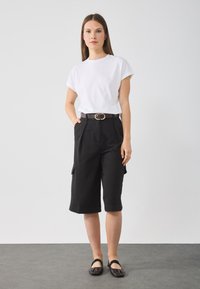 T-shirt blanc à manches courtes, bermuda cargo noir avec poches latérales, ballerines noires et une ceinture marron avec une boucle circulaire. Couleurs unies et design classique.
