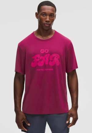 Uomo che indossa una maglietta magenta con il testo "GO FOR FOR ALL RUNNERS", abbinata a pantaloncini scuri, in piedi davanti a uno sfondo chiaro e uniforme.