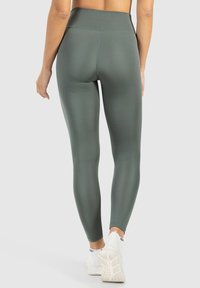 Vysoké olivově zelené legíny vyrobené z elastického materiálu; hladký povrch s plochými švy, kombinované s bílými sportovními botami.