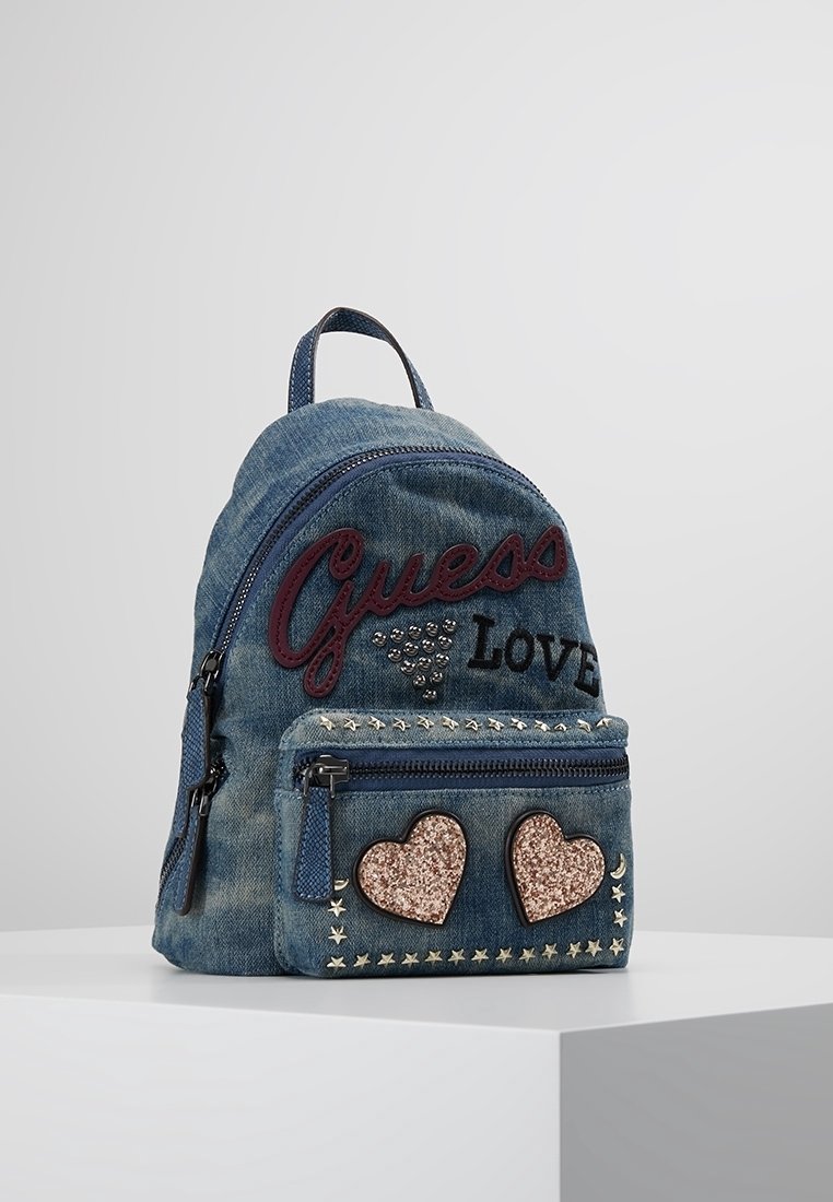 Sac à dos en jean avec texte "Guess Love", clous métalliques et patches en forme de cœur scintillants sur la poche avant, posé sur une surface blanche.