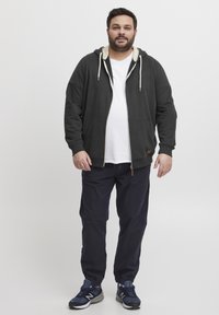 Felpa con zip di colore grigio scuro con dettagli crema, abbinata a pantaloni blu navy e sneaker blu navy. Presenta tasche a marsupio e cappuccio con cordino.
