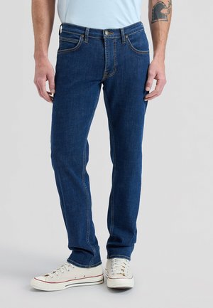 Homme portant un jean droit bleu foncé, des baskets blanches et une chemise bleu clair, debout les mains détendues le long du corps.