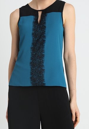 Mouwloze teal top met zwart juk en verticale zwarte kanten bies in het midden, gecombineerd met zwarte broek.