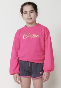 Un sweat-shirt long en rose avec le texte doré "Blossom", associé à un short gris avec des bords roses et un petit logo en cœur.