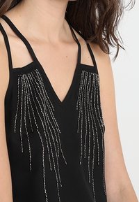 Black sleeveless top with a V-neck and thin straps, decorated with silver beaded fringe detailing cascading from the chest.