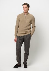 Beige gerippter Pullover mit einem Viertelreißverschluss und einer Brusttasche, kombiniert mit dunkelgrauen Hosen und braunen Slippern.
