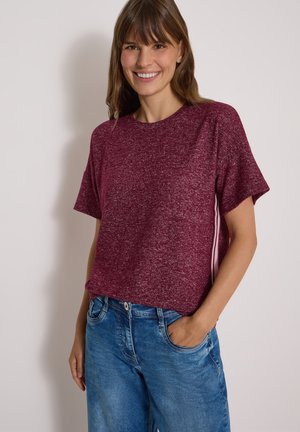 Jeune femme souriante portant un t-shirt bordeaux à manches courtes et un jean bleu, une main dans la poche, sur un fond clair uni.