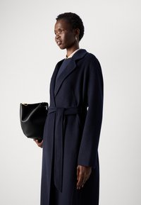 Mulher com cabelo curto, vestindo um casaco azul marinho com cinto e segurando uma bolsa preta, de pé diante de um fundo claro e liso.