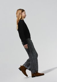 Suéter negro de punto, pantalones con estampado de lentejuelas en negro y plateado, y botines marrones de ante con detalle de herrajes laterales. Modelo en pose de perfil.