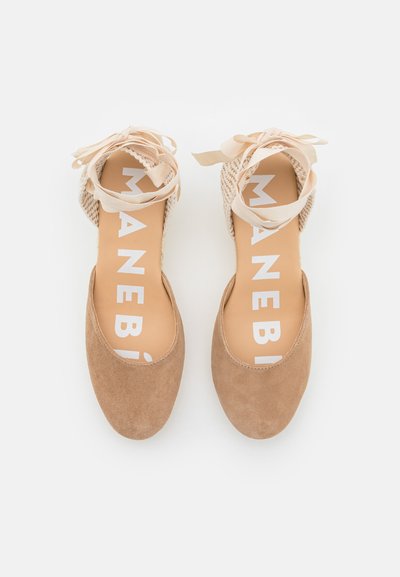 Scarpe basse in suede tan con dettagli intrecciati beige e un nastro legato alla caviglia. La soletta presenta un marchio bianco su uno sfondo chiaro.