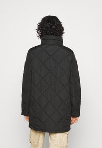 Svart quiltad kappa med hög krage, med ett diamantmönster och långa ärmar, visad från baksidan med en lätt rundad fåll.