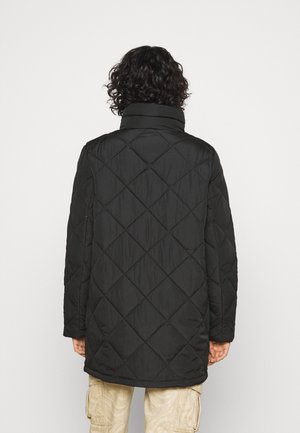 Svart quiltad kappa med hög krage, med ett diamantmönster och långa ärmar, visad från baksidan med en lätt rundad fåll.