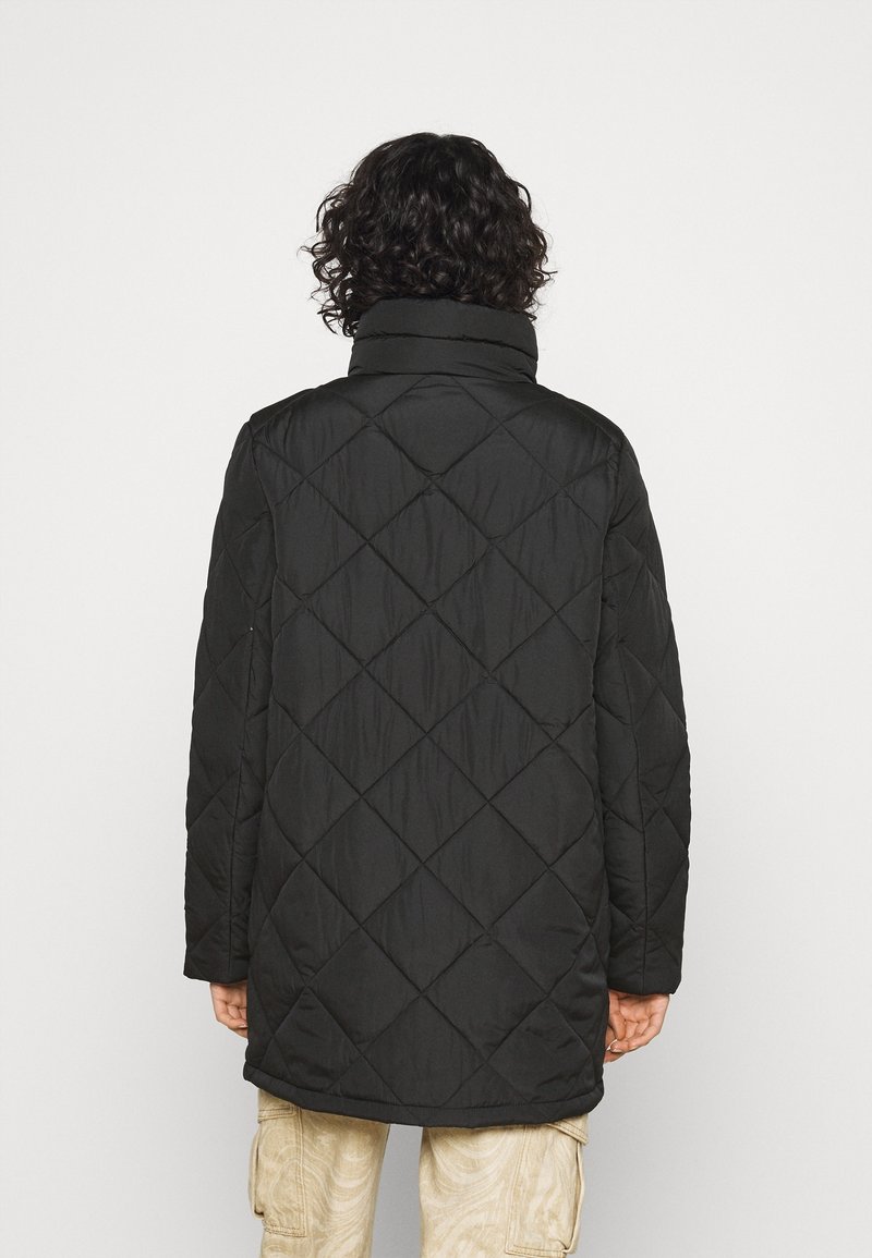 Svart quiltad kappa med hög krage, med ett diamantmönster och långa ärmar, visad från baksidan med en lätt rundad fåll.