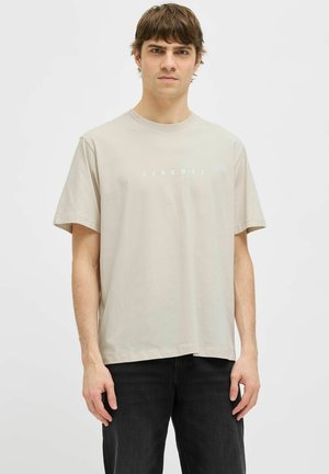 Joven con cabello castaño corto que lleva una camiseta beige de JJ Rebel y pantalones negros sobre un fondo blanco liso.