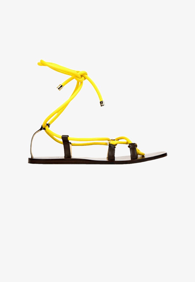 Sandale plate avec une fine semelle noire, des lanières noires et des lacets en corde jaune vif noués autour de la cheville.