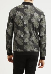 Uomo che indossa una maglietta nera a maniche lunghe con un motivo di foglie tropicali verdi e fiori bianchi, in piedi con la schiena rivolta verso uno sfondo semplice.