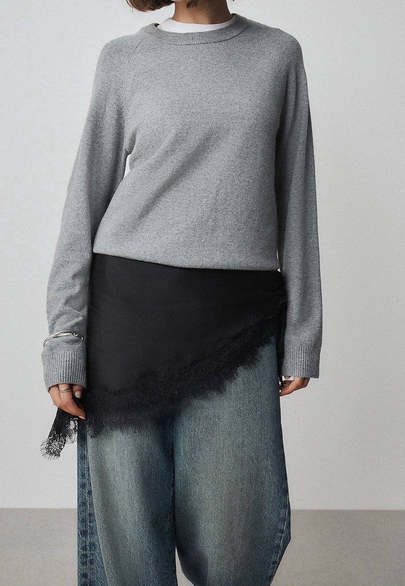 Personne portant un pull gris clair à manches longues sur un jean bleu à jambes larges et une jupe asymétrique noire avec bordure en dentelle.