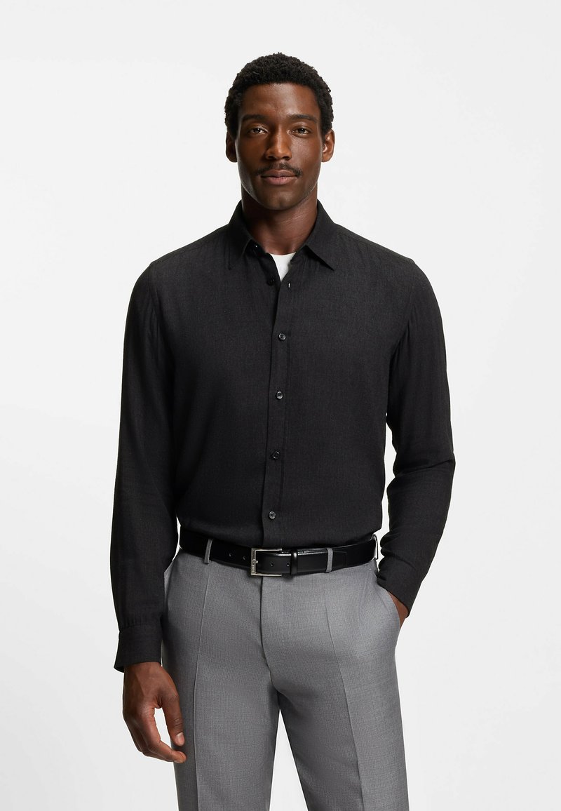 Camicia nera a maniche lunghe con bottoni, dal tessuto liscio e colletto a punta, abbinata a pantaloni grigi chiari e una cintura nera con fibbia argentata.