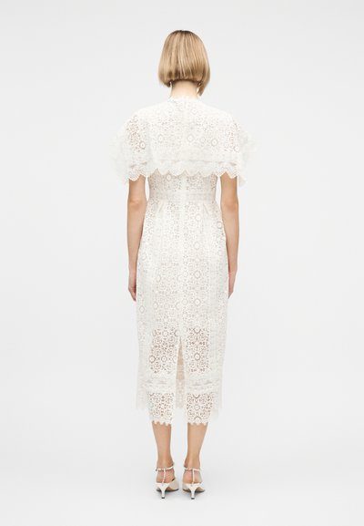 Γυναίκα με κοντά ξανθά μαλλιά φοράει λευκό δαντελένιο μίντι φόρεμα και λευκά σανδάλια με τακούνι, στέκεται αντικρίζοντας ένα απλό ανοιχτό γκρι φόντο.