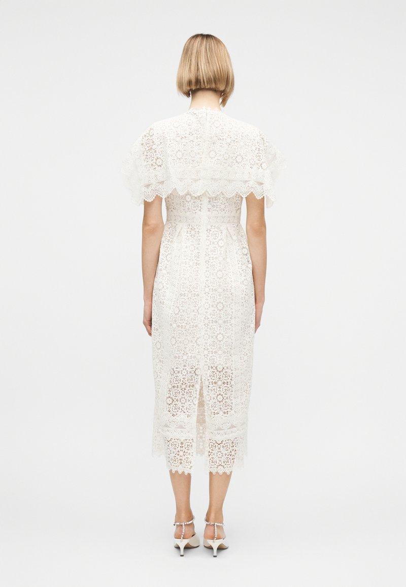 Frau mit kurzem blondem Haar trägt ein weißes Spitzen-Midikleid und weiße Sandaletten mit Absatz, sie steht vor einem schlichten hellgrauen Hintergrund.