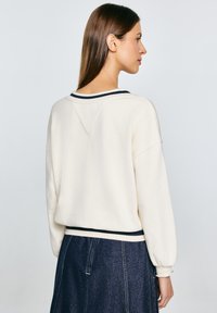 Cremfarbener Pullover mit einem V-förmigen Rückendesign, schmalem schwarzen Streifen am Kragen und lässigem Schnitt. Kombiniert mit einem Jeansrock.