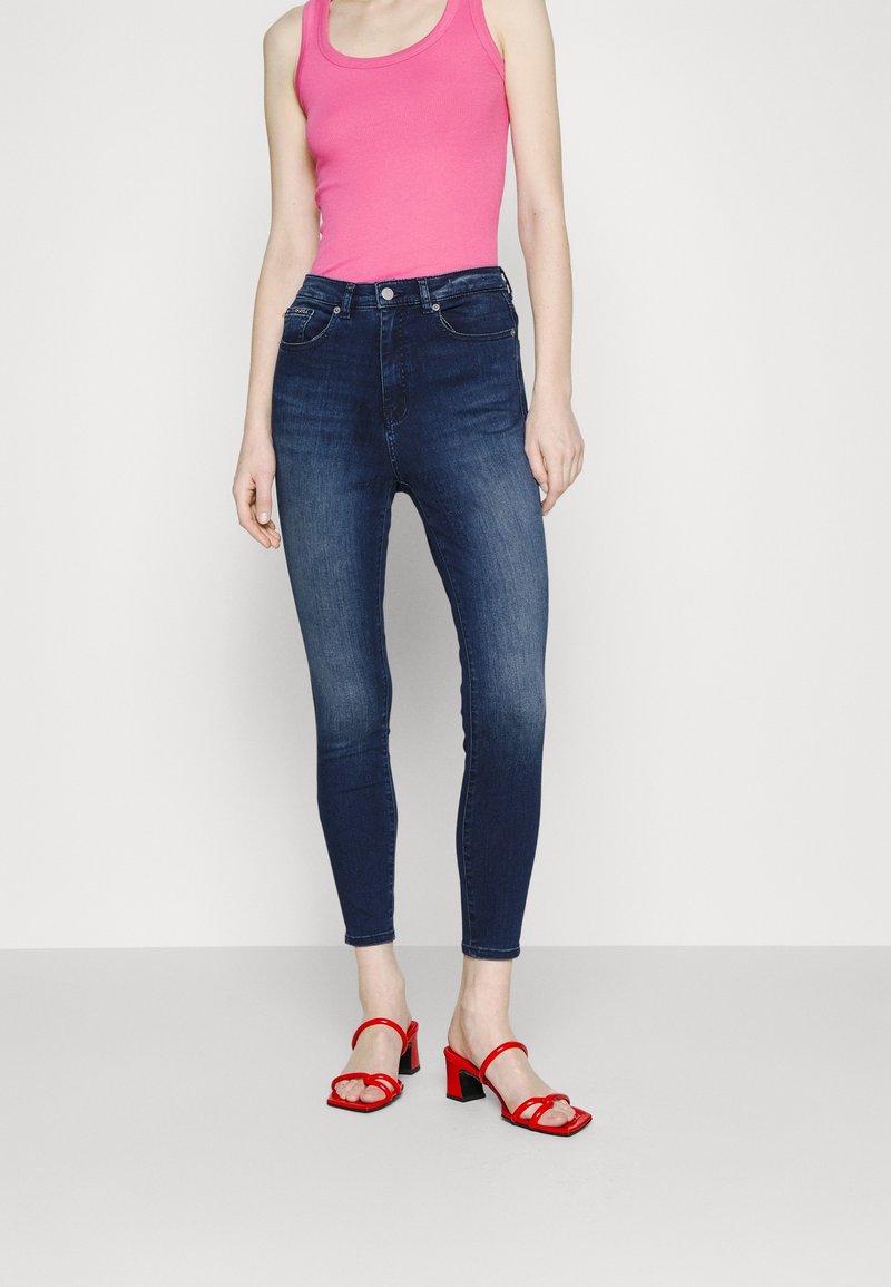 Jeans de tiro alto de color azul oscuro combinados con una camiseta ajustada rosa y sandalias de tacón rojo. Textura de denim suave con bolsillos frontales.