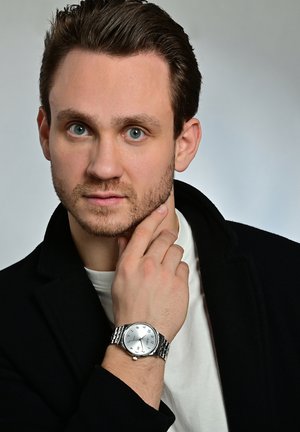 Silberne Edelstahl-Armbanduhr mit rundem Ziffernblatt, klaren Ziffernmarkierungen und einem Metallarmband, getragen von einer Person in nachdenklicher Pose.