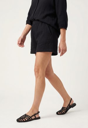 Kvinde går iført sorte teksturerede shorts, sorte vævede slingback flade sandaler og en sort top med lange ærmer mod en ensfarvet baggrund.