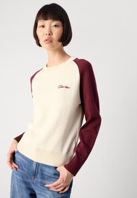 Young person with short dark hair wears cream and burgundy long-sleeve sweater and blue jeans, standing against plain light background.