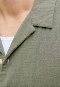 Olijf groene linnen shirt met een kraag, knoopsluiting en gestructureerde stof. Witte onderhemd zichtbaar onder de kraag.