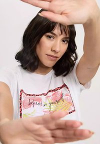 Biała koszulka z prostokątną grafiką przedstawiającą różowe i zielone liście tropikalne, osadzoną w leopardzim obramowaniu, z napisem "uwierz w siebie."