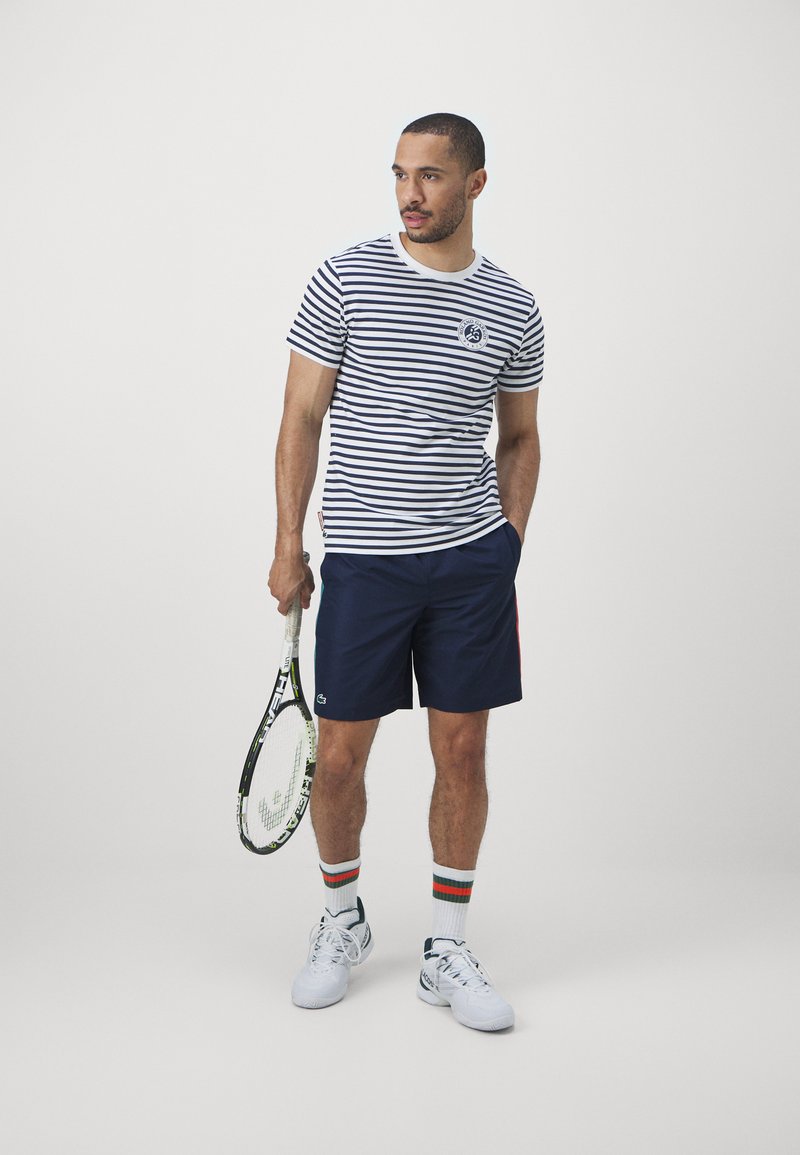 Chemise à manches courtes rayée bleu marine et blanche, assortie à un short bleu marine. Il tient une raquette de tennis, portant des chaussures blanches et des chaussettes rayées.