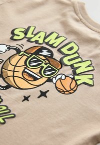 T-shirt color beige con una grafica di un pallone da basket con occhiali da sole, testo che recita "SLAM DUNK" e stelle con accenti verdi e neri.