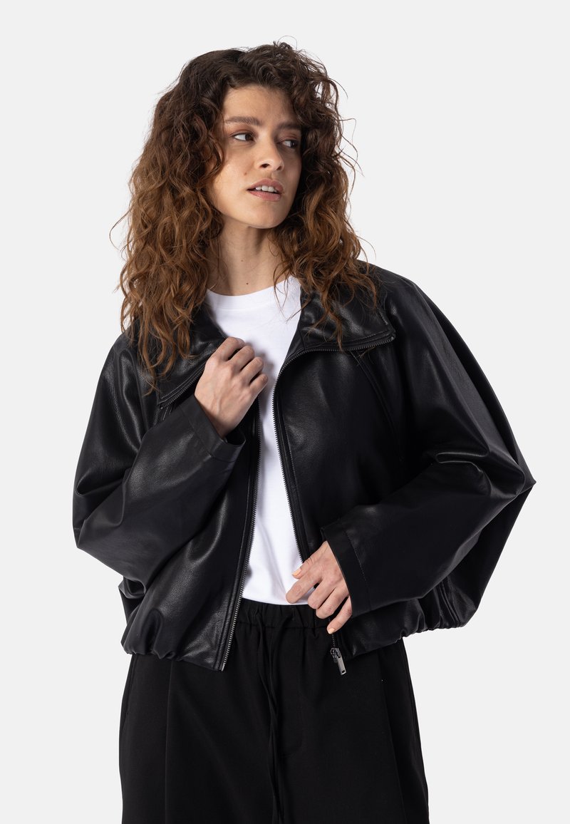 Femme aux cheveux bouclés portant une veste en cuir noire par-dessus une chemise blanche et un pantalon noir, regardant sur le côté sur un fond clair uni.