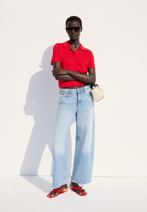Osoba nosiaca červené polo s krátkym rukávom, svetlomodré rifle s širokými nohavicami, červené sandále, slnečné okuliare a drží malú bielu kabelku.