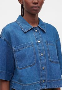 Woman wearing a blue denim short-sleeve shirt with large front pockets and silver buttons, shown from shoulders to waist.